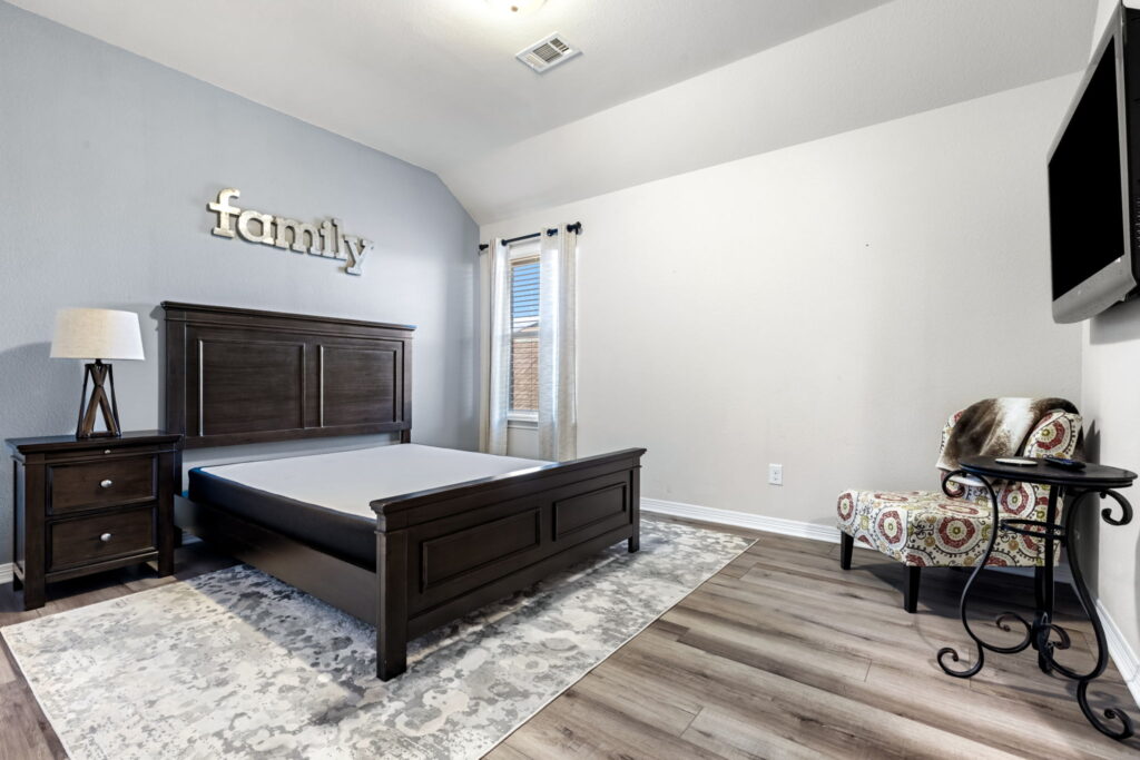 A spacious bedroom with light gray walls, dark wood furniture, wood-look flooring, and a patterned accent chair in the corner.